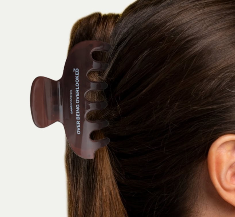 A brown hair clip holds back brown hair. The clip is printed with the words "OVER BEING OVERLOOKED." The image shows the side and back of a person's head.