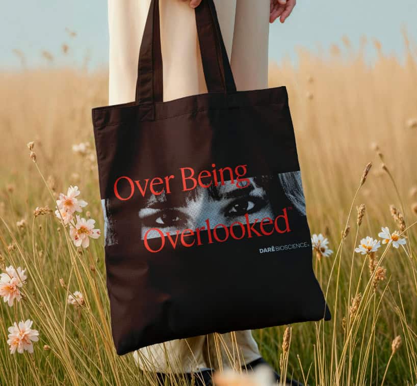 A person standing in a field holds a black tote bag with an image of eyes and the text "Over Being Overlooked" in red.