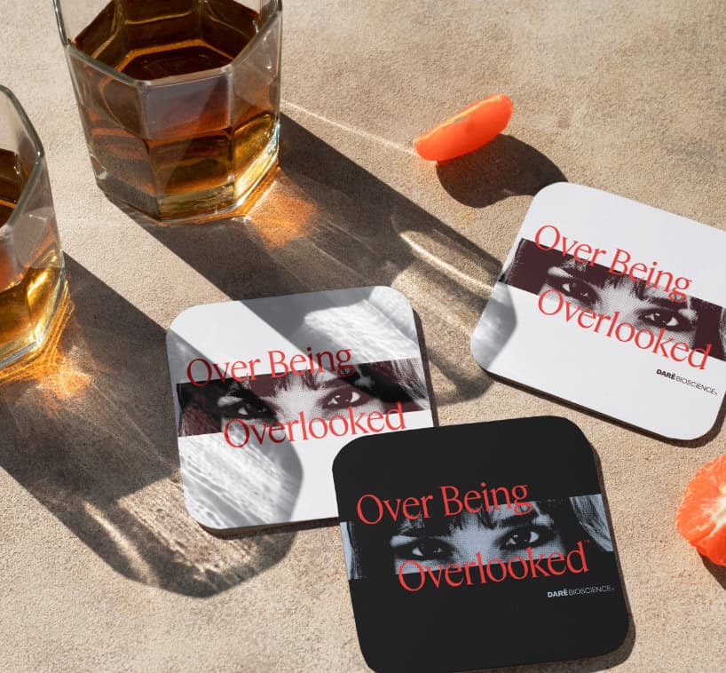 Two glasses of whiskey and three coasters with an image of eyes and the text "Over Being Overlooked" on a sunlit surface; an orange peel is nearby.