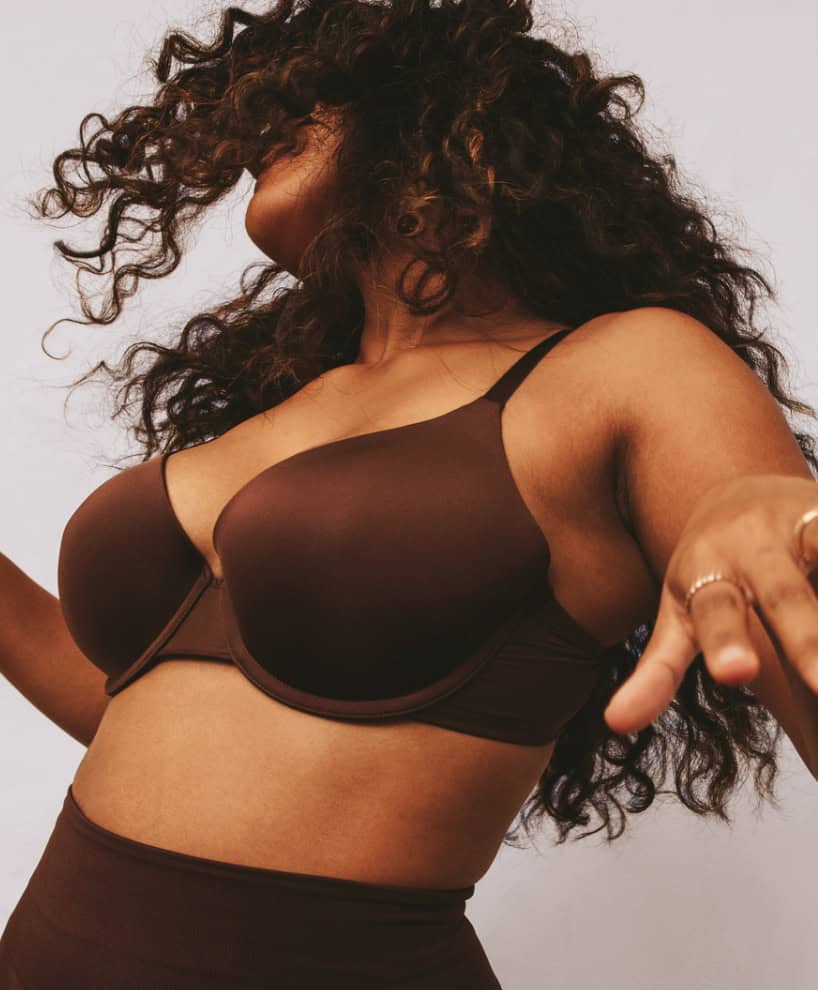 A person with long curly hair wearing a dark brown bra and high-waisted bottoms stands against a neutral background, mid-movement, with arms outstretched.