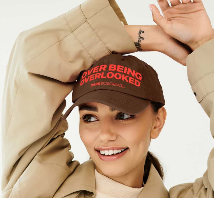 Woman in beige trench coat and brown cap with red text that reads "OVER BEING OVERLOOKED" and "DARE BIOSCIENCE," smiling and posing with arms raised.