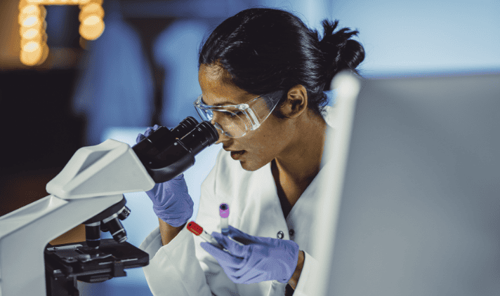 A scientist wearing safety goggles and gloves examines a sample slide under a microscope in a laboratory setting.