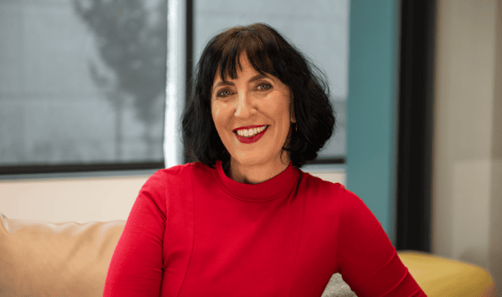 Woman with short black hair wearing a red top is sitting indoors, smiling at the camera. A window and blue wall are visible in the background.